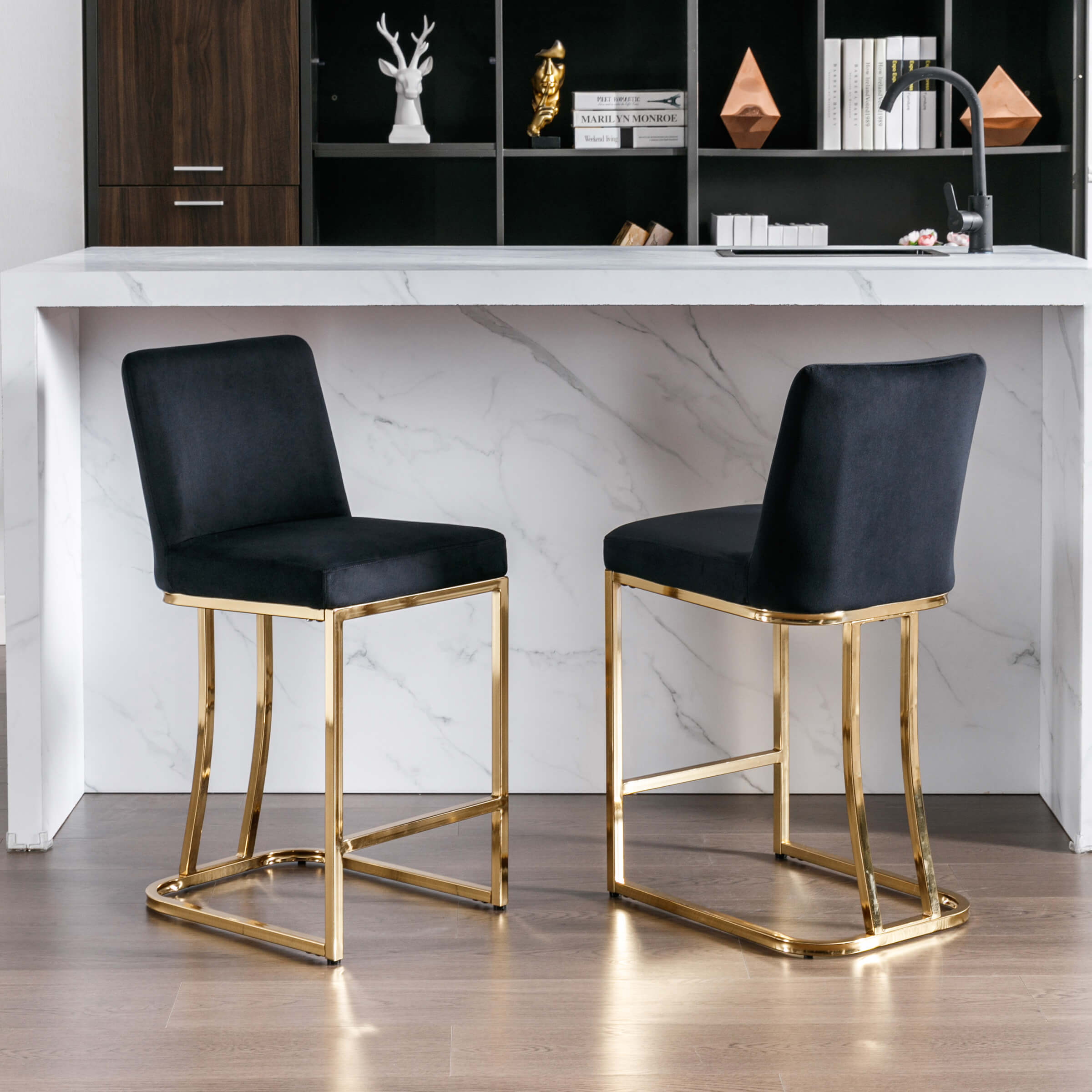 Two bar stools with velvet upholstery and gold frames displayed in front of shelves